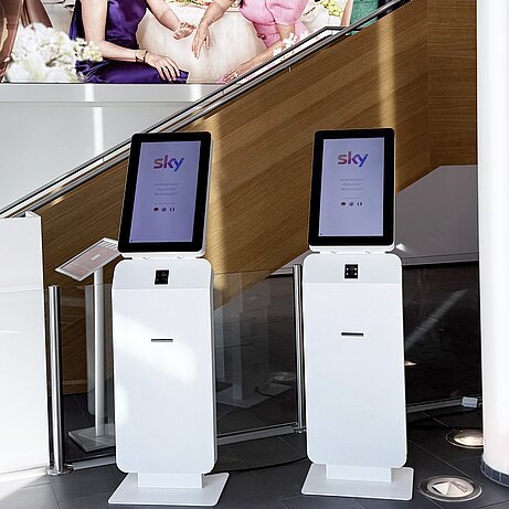 Zwei CheckBox-Terminals im Foyer eines Sky-Standorts, dahinter eine Treppe und eine Werbewand.