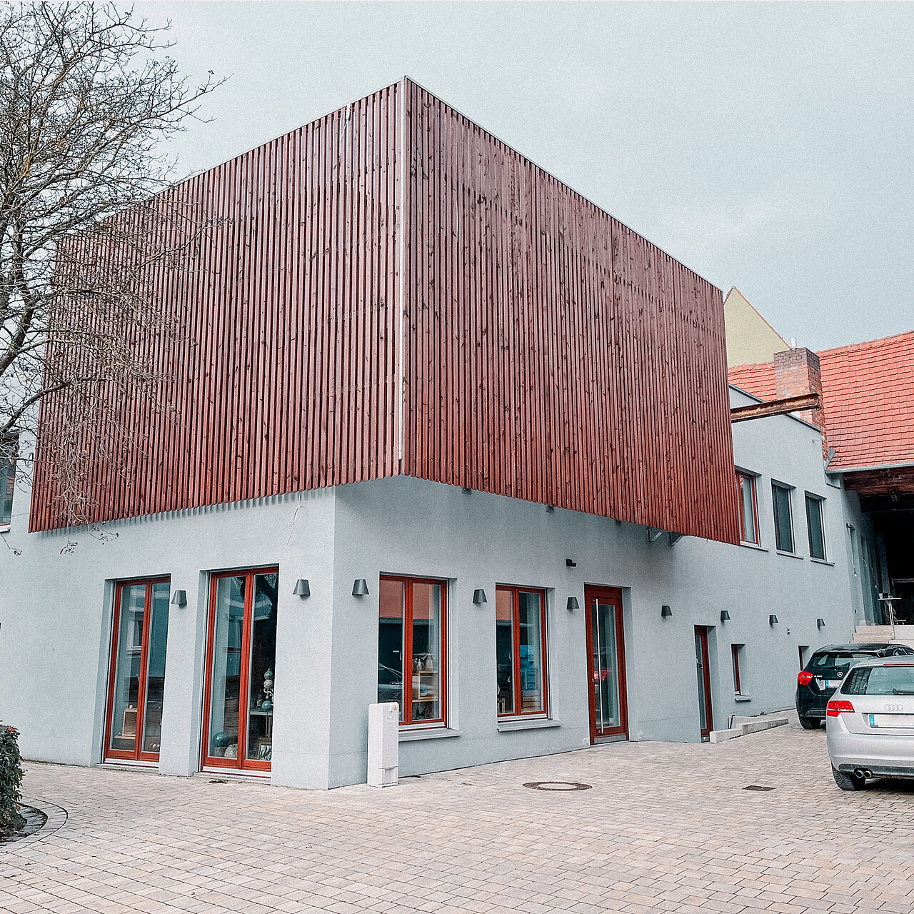 Zweistöckiges Firmengebäude mit grauem Erdgeschoss und Holzverkleidung im Obergeschoss, roten Fensterrahmen und geparkten Autos davor; moderne Architektur neben traditionellen Häusern mit Ziegeldach.